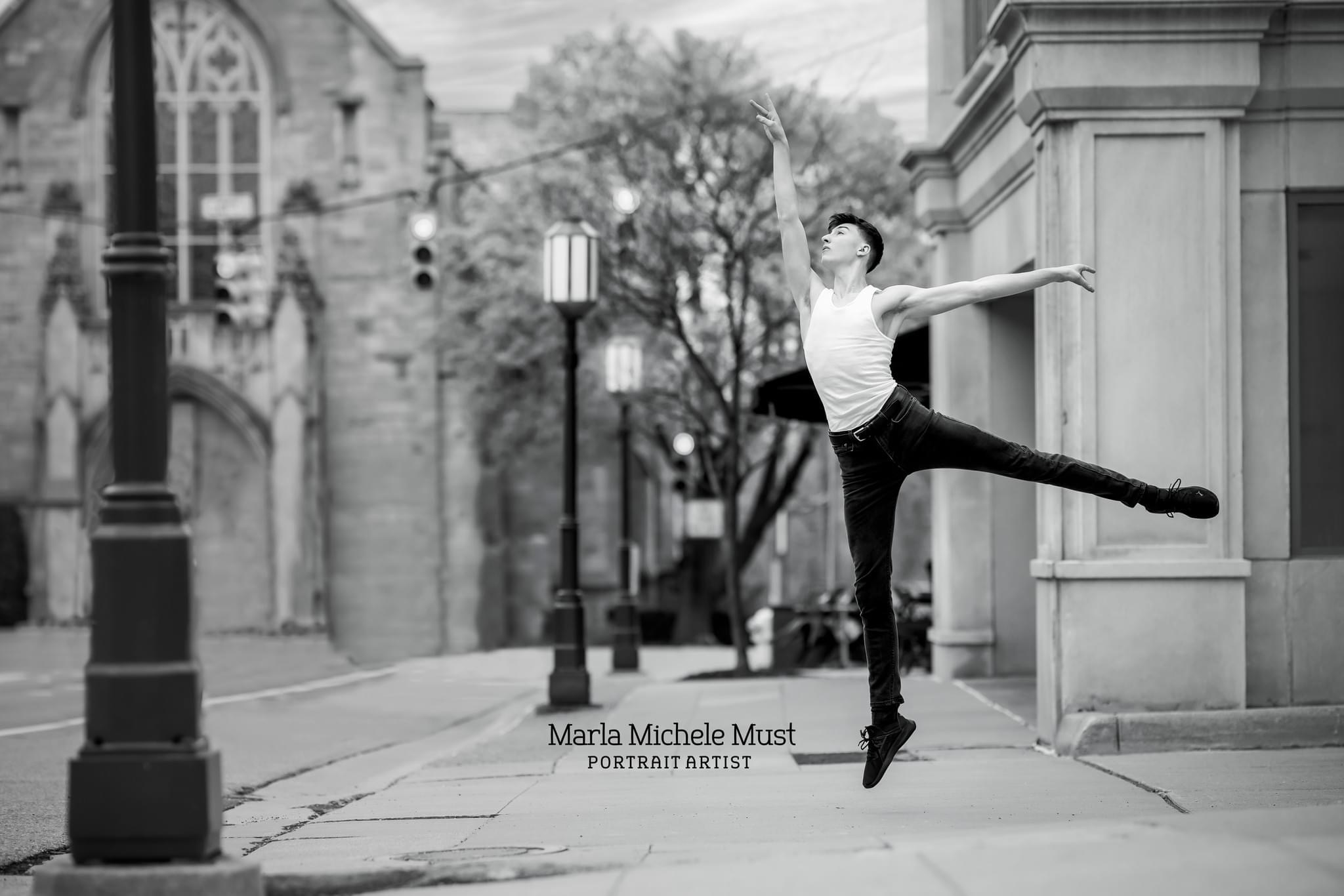Colorless photo of a man posing with pointed toes and an arm outstretched above him along a lane of Detroit city streetlamps during a dance photoshoot, taken by a Michigan dance photographer.