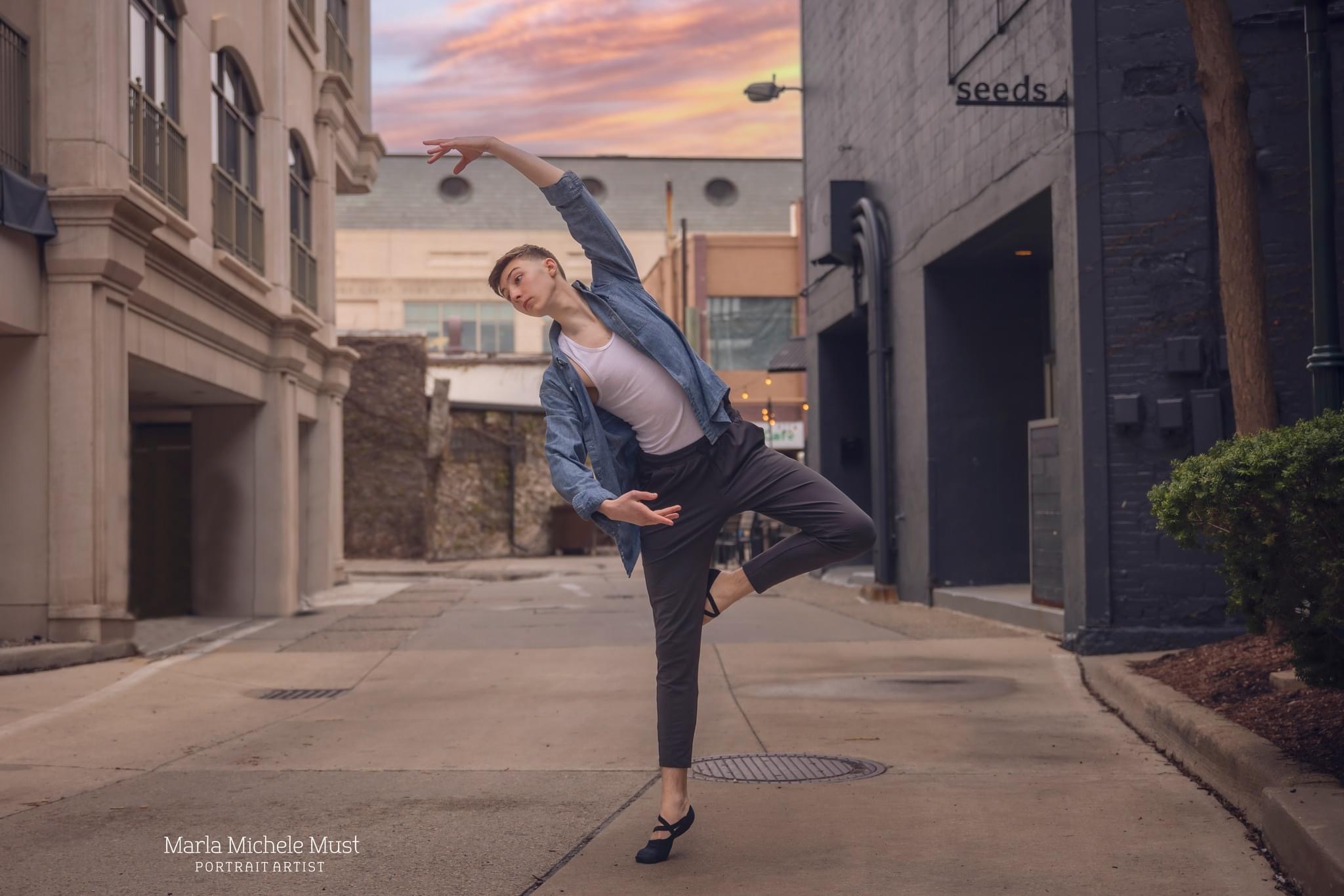 Man in casual clothing poses in a city with one arm above his head and one at his waist during a dance photoshoot, taken by a Michigan dance photographer.