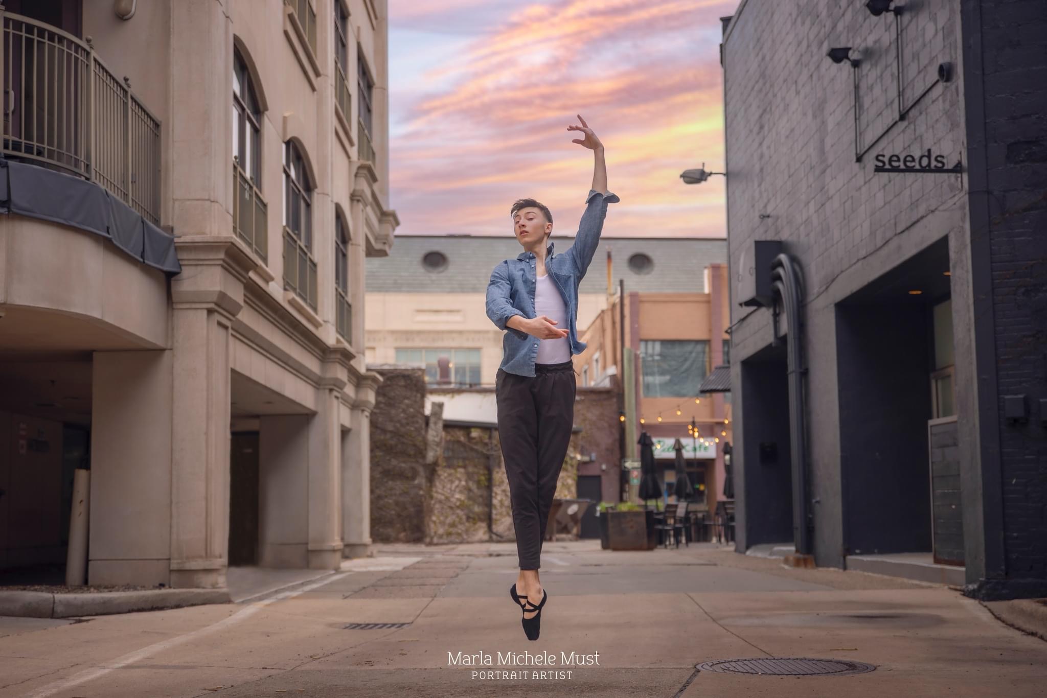 Man poses on pointed toes with one arm outstretched above his head in casual clothing during a dance photoshoot in the city, taken by a Michigan dance photographer.