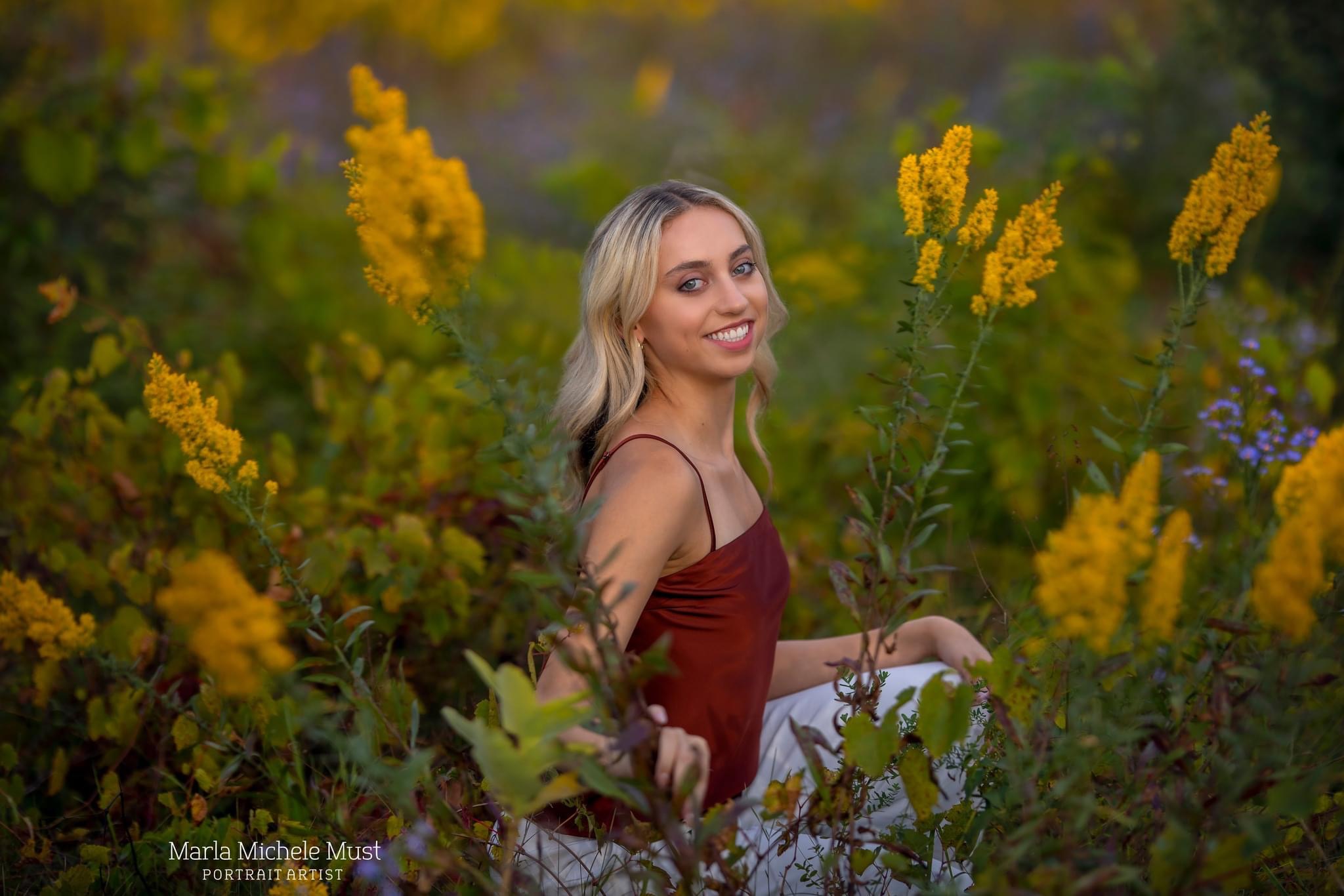 A high school senior poses for her senior photoshoot while sitting among a field of yellow flowers