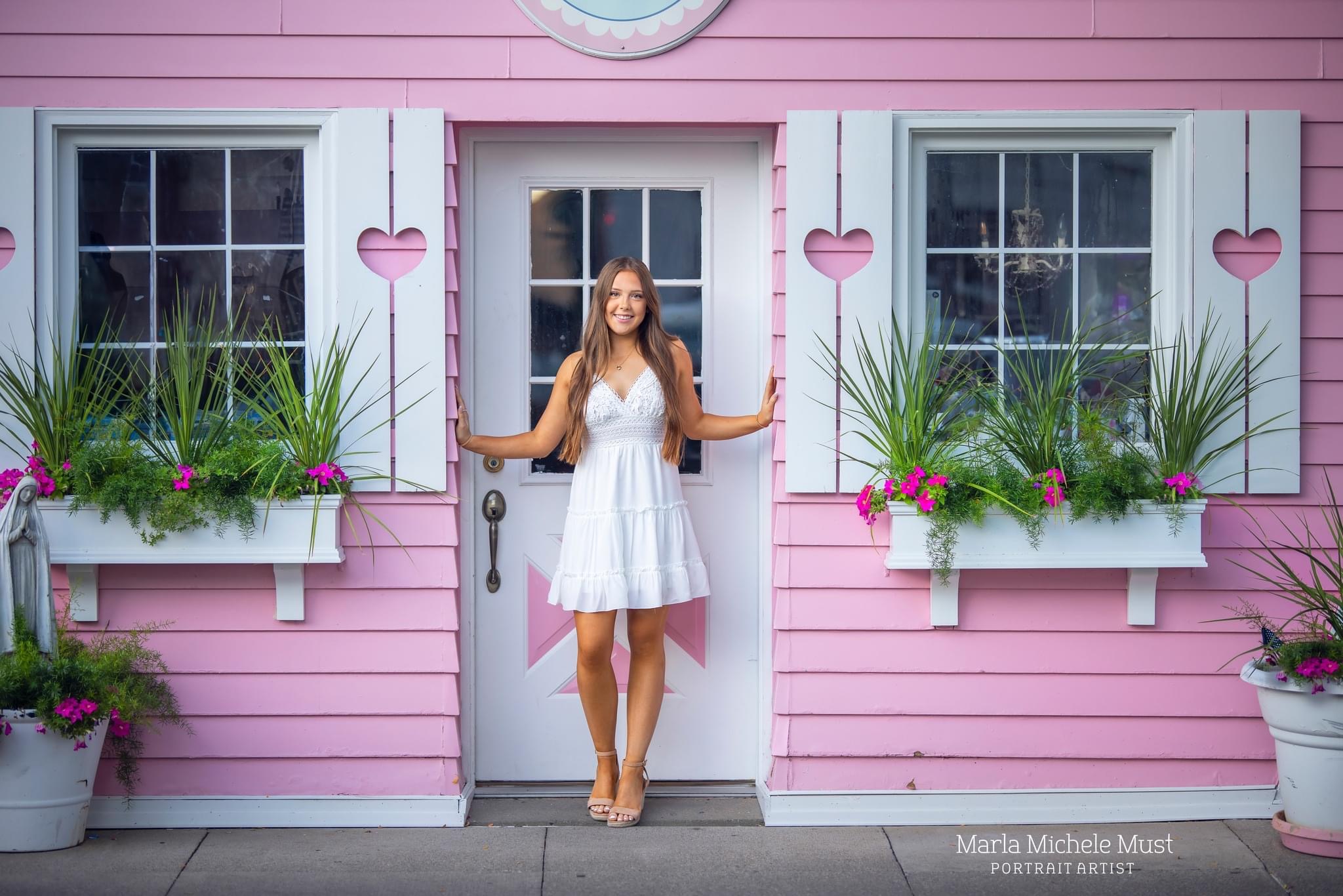 A high school senior poses for her graduation photo while standing in the doorway of a pink house and flower beds within a Detroit neighborhood