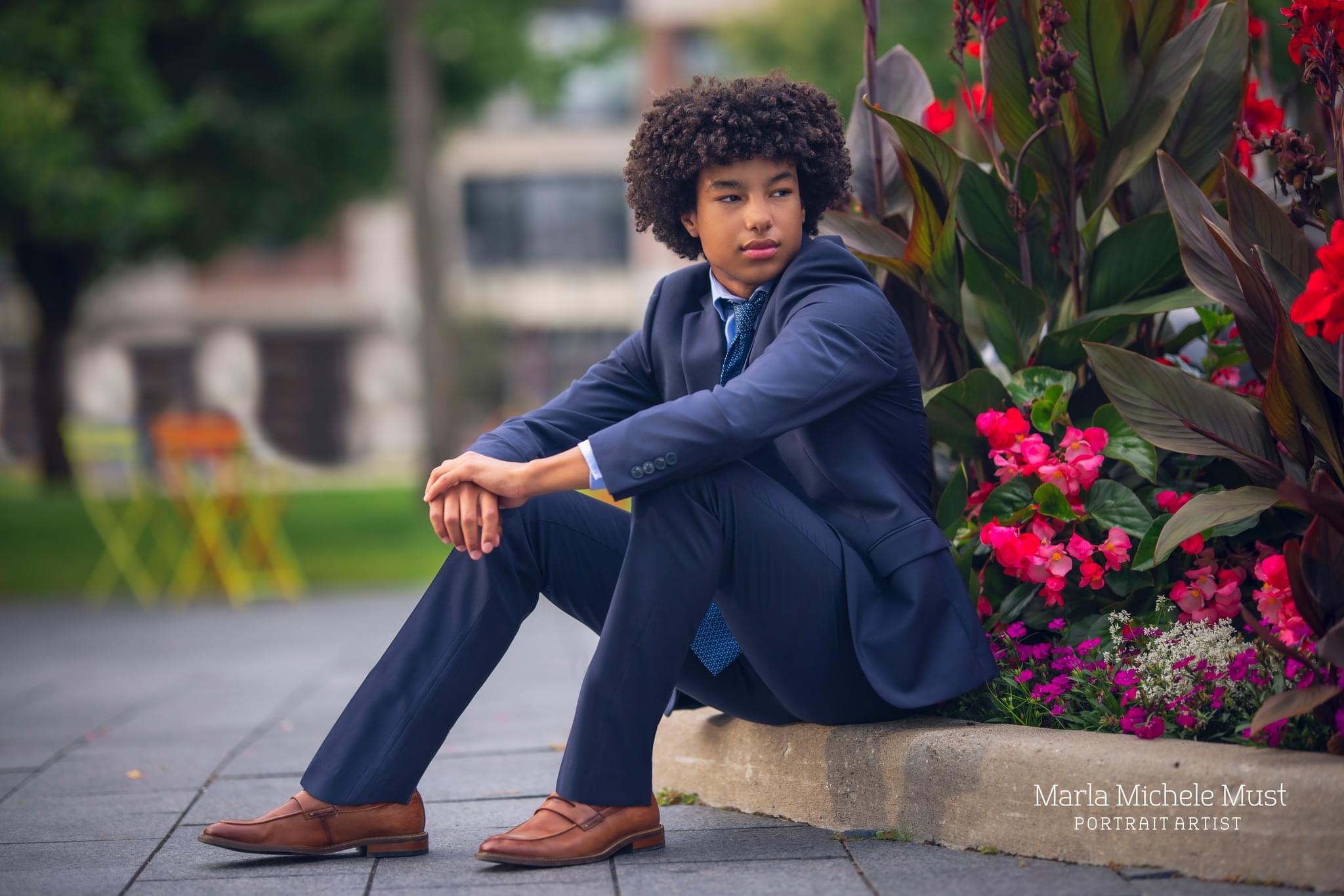 A young man poses for his senior photoshoot in a formal suit while sitting on the edge of a downtown Detroit flower bed