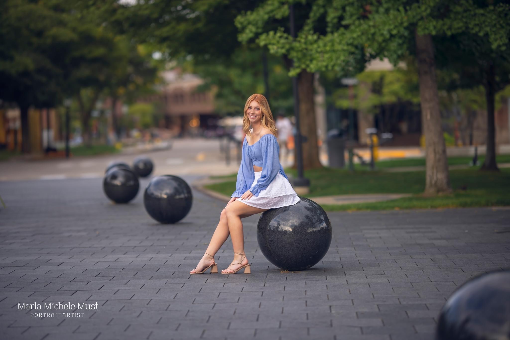 A young woman poses for her senior photoshoot while sitting on a spherical sculpture in a Downtown Detroit park