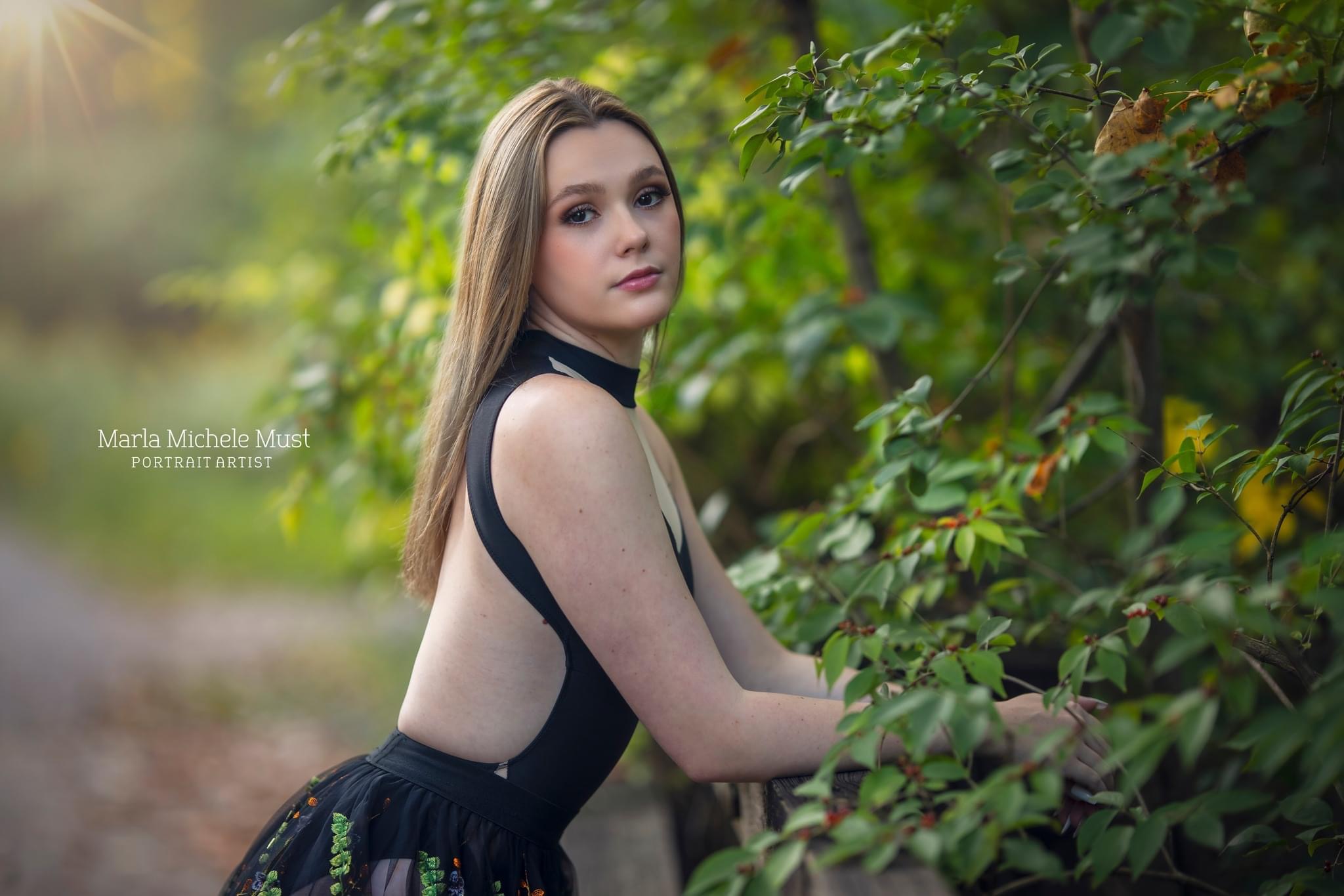 A young woman poses for her senior portrait in a classy black dress as she leans against a Detroit garden hedge