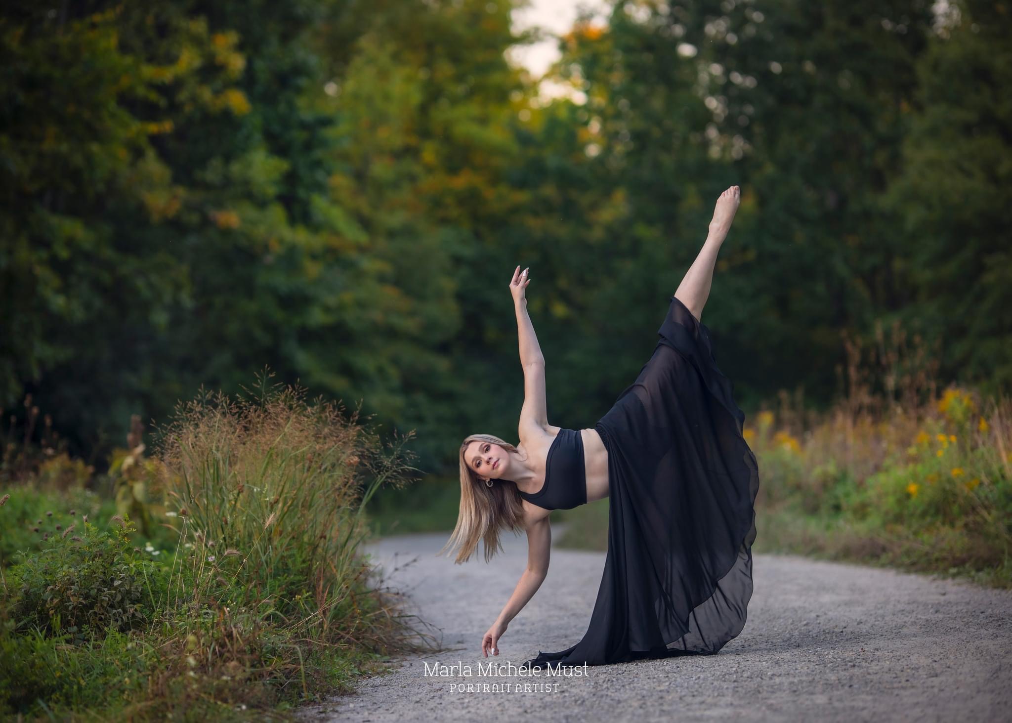 Dancer in a flowy black dress poses in a leaning pose on a Detroit park pathway for a dance photoshoot.