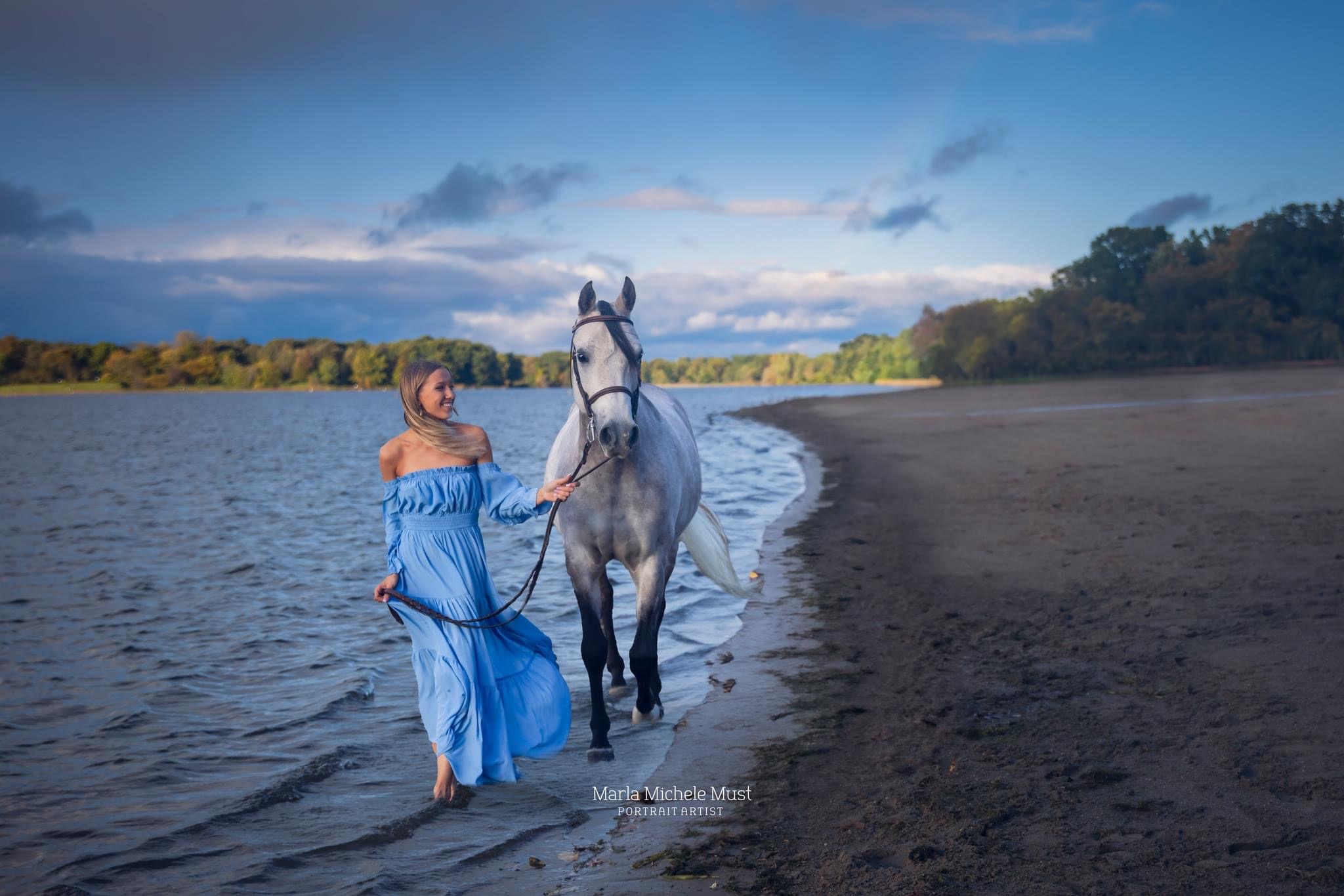 A girl in a blue dress walks along a Michigan beach with her horse for her high school senior pictures
