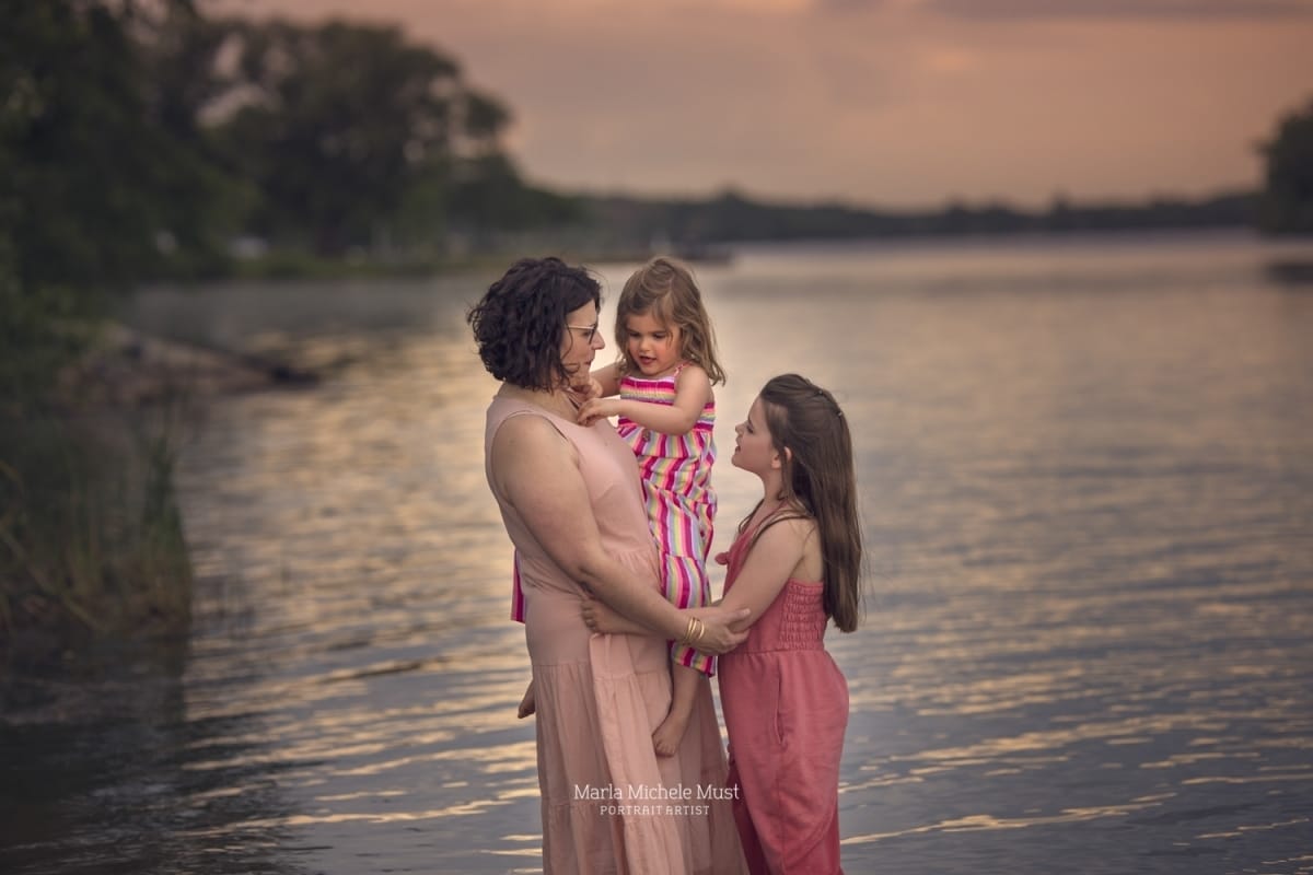 A family of a mother and two daughters wear pink and pose for a family photo on a shoreline in Michigan.