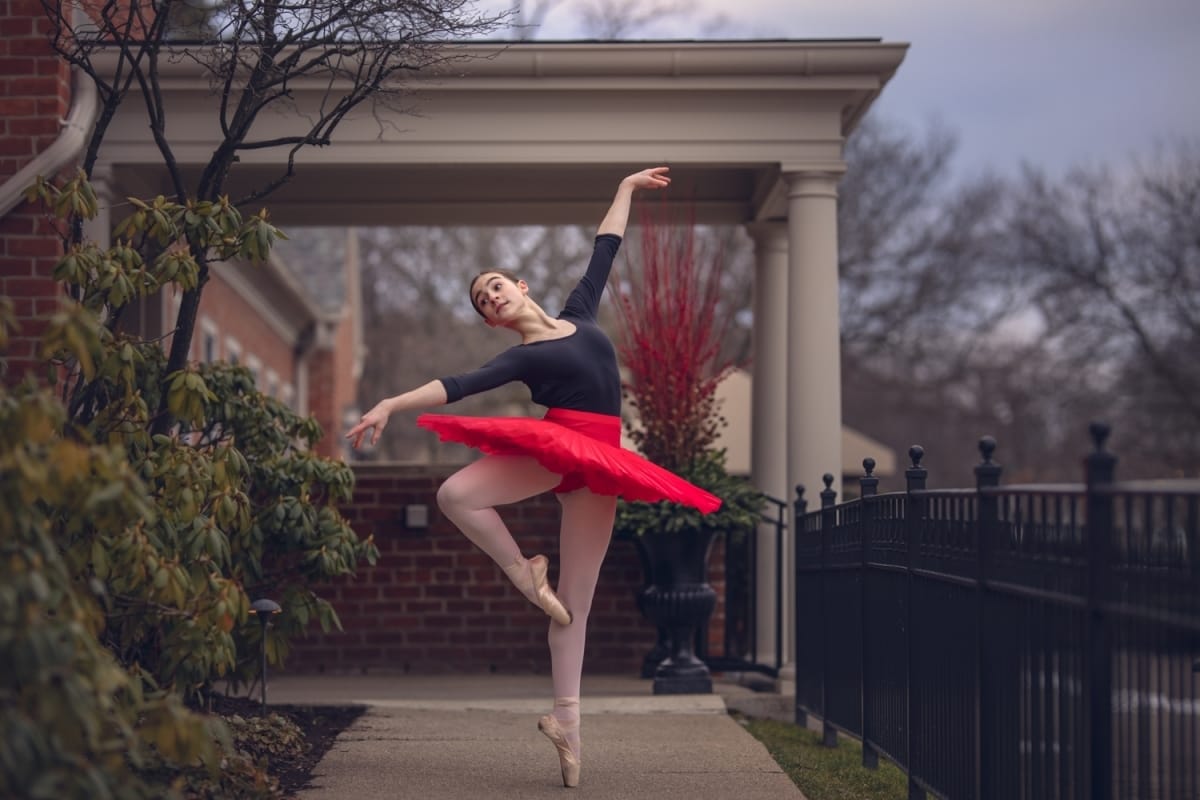 Michigan ballerina poses gracefully with arms extended and one leg slightly bent as she dances outside for a Detroit dancing photoshoot.