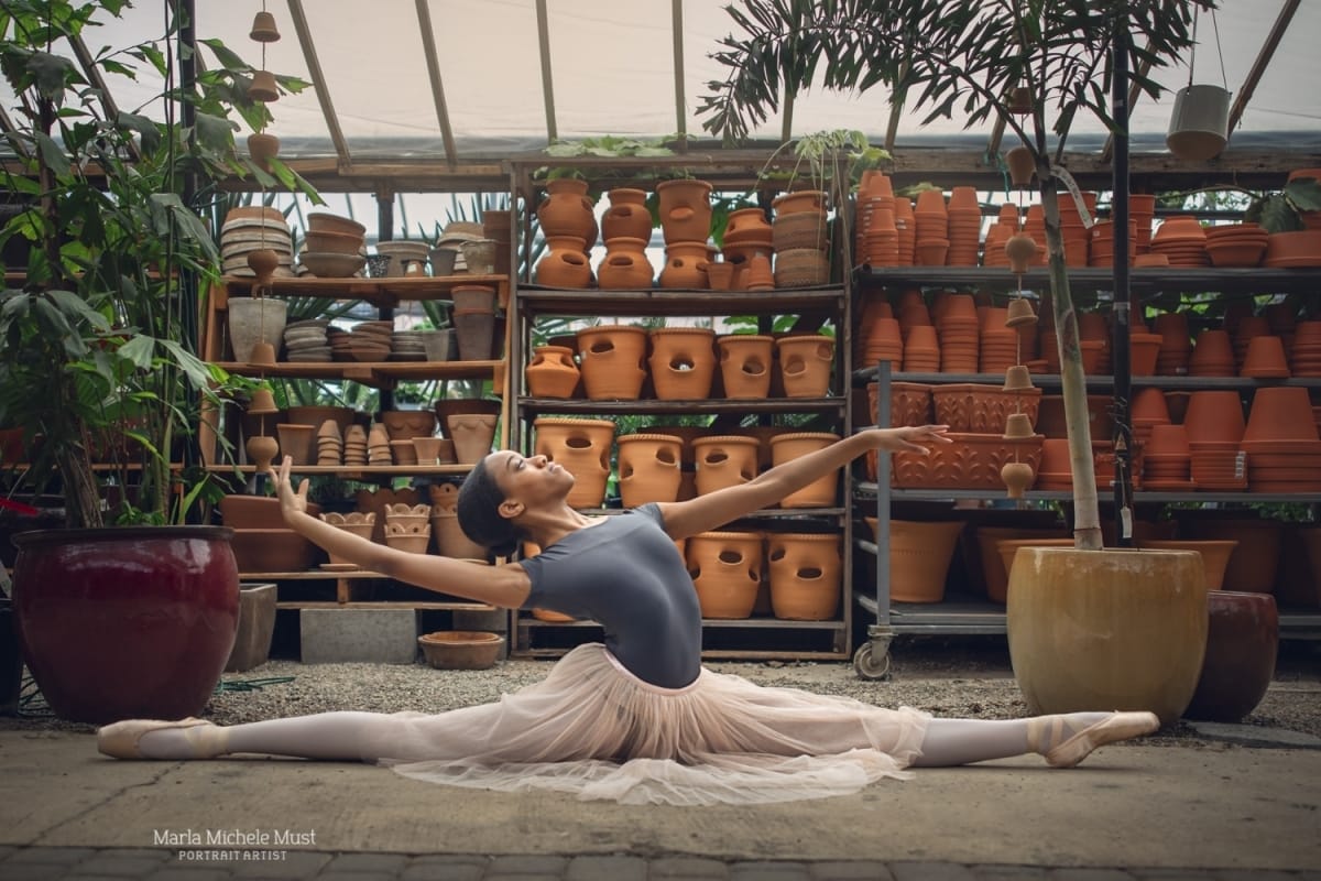 Dancer, wearing a cream-colored tutu and grey leotard, strikes a beautiful pose in the splits, beautifully photographed in a Michigan flower nursery by a skilled Detroit dance photographer.