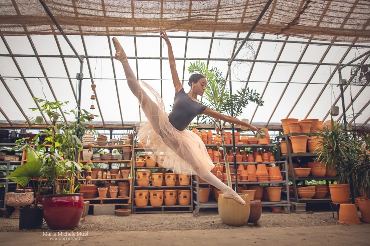Dancer, wearing a cream-colored tutu and grey leotard, strikes a beautiful pose in the air, beautifully photographed in a Michigan flower nursery by a skilled Detroit dance photographer.