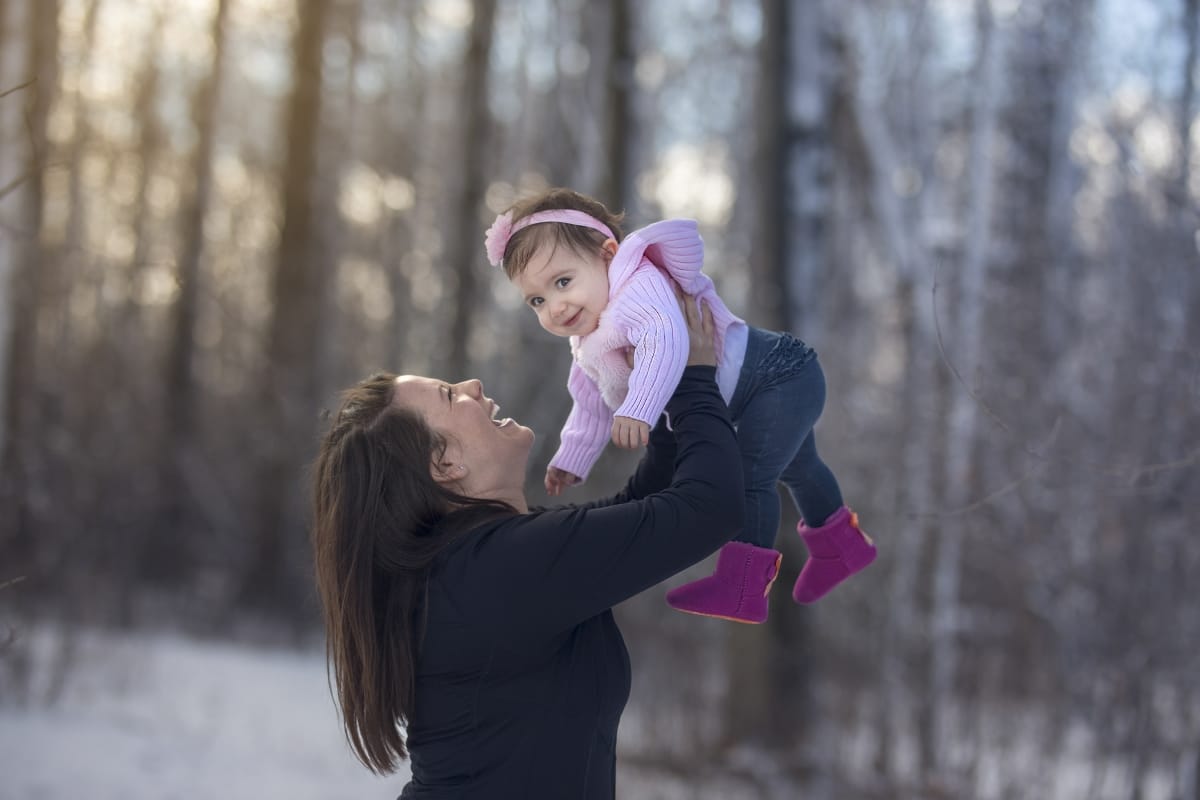A mother holds up her young baby above her head while laughing. A snowy landscape is behind her.