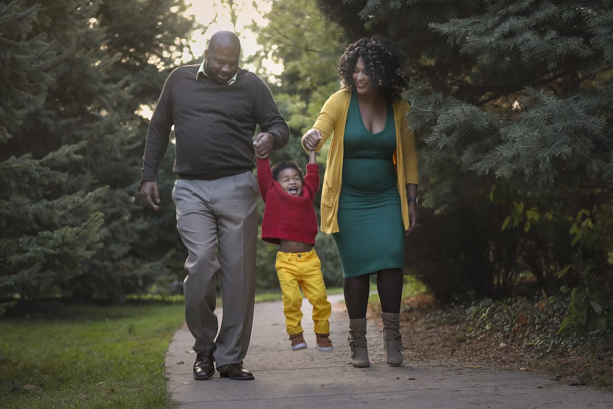 A portrait of a couple holding hands with their young child and lift them in the air while walking along a park trail in Detroit.