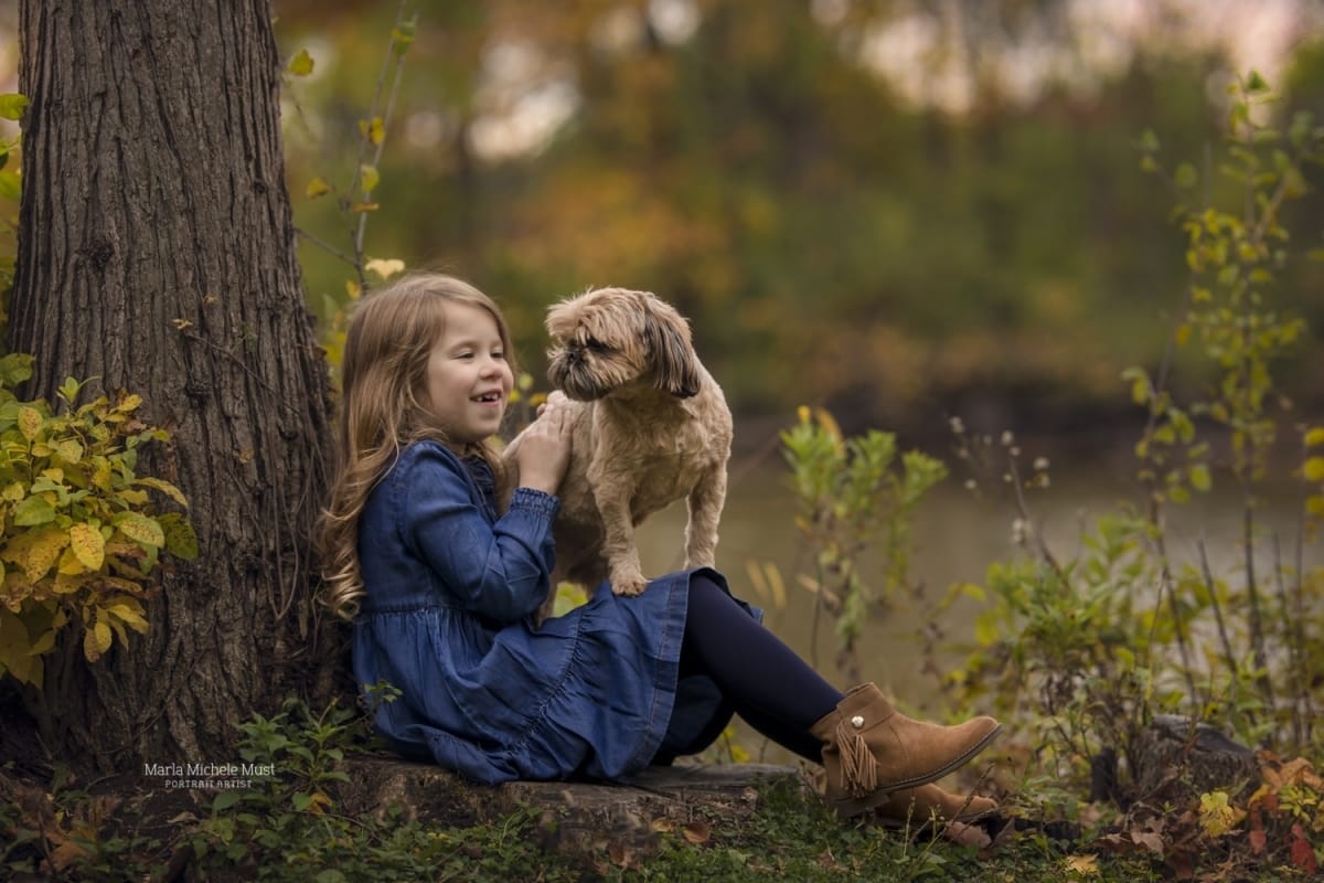 A young girl sits with her back to a tree while a small dog stands on her lap.