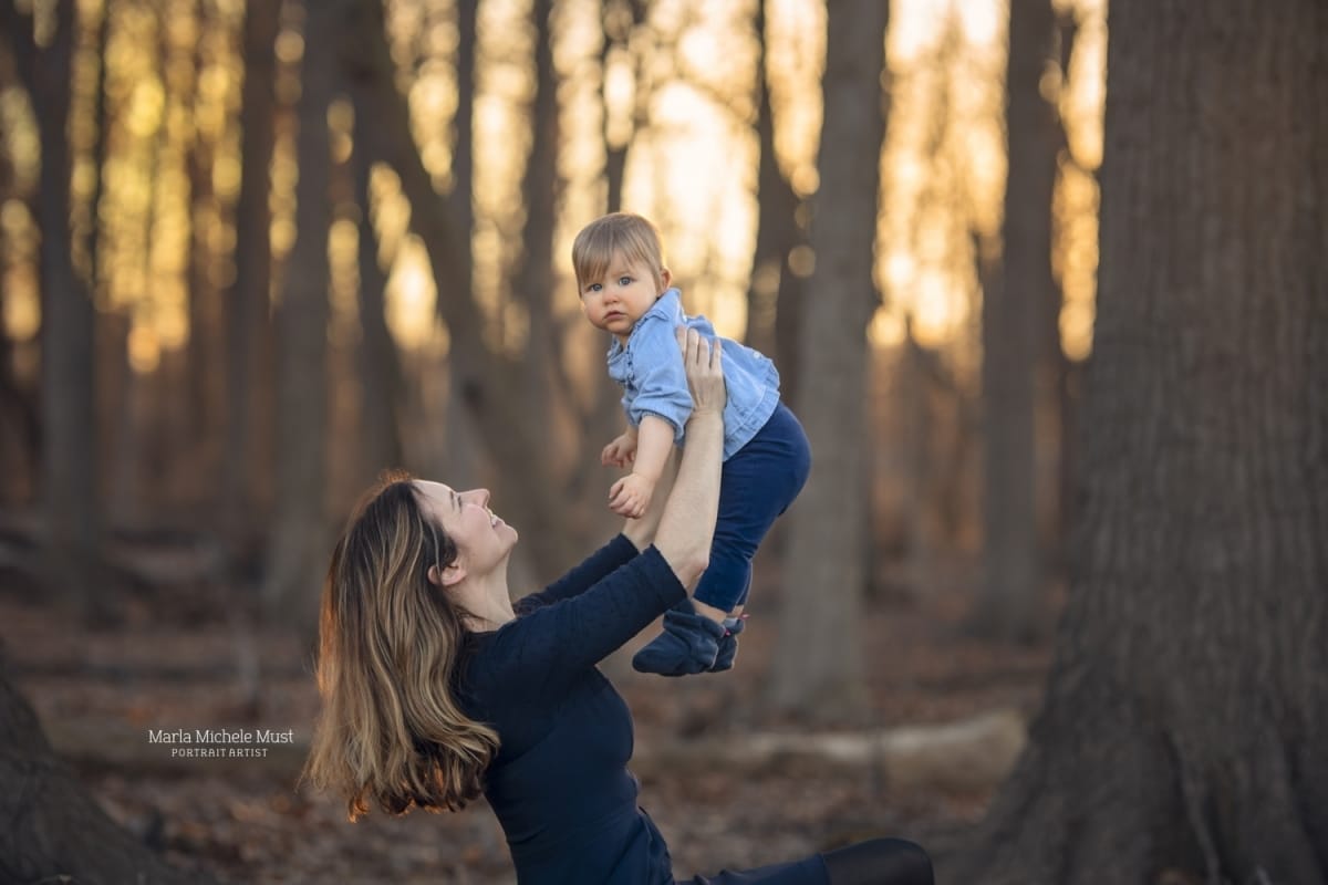 A photo of a woman lifting her baby in the air while smiling in a Detroit forest during sunset.