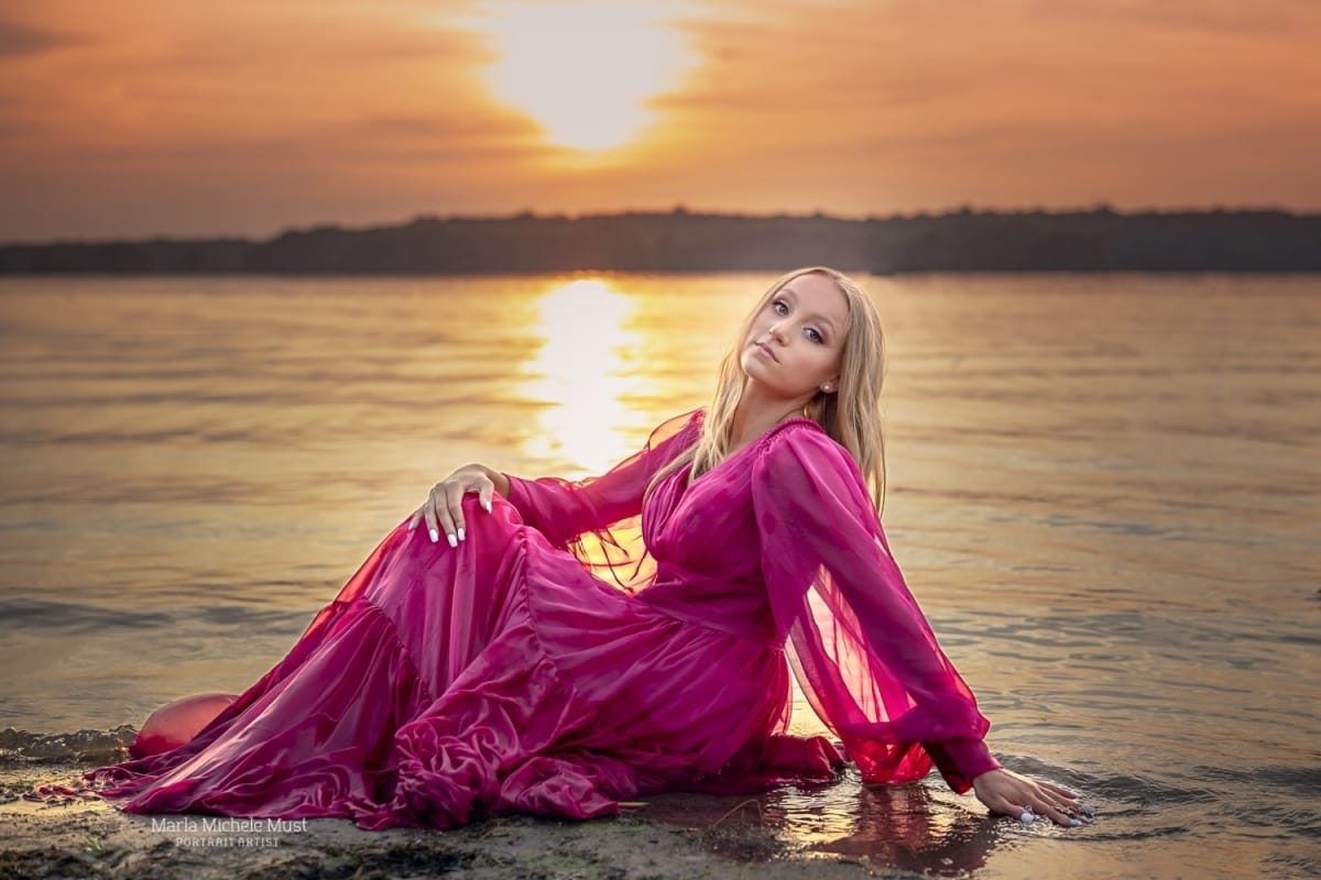 Romantic senior photo taken by a skilled Detroit photographer depicting a blonde girl in a long fuchsia gown sitting on the shallow waters of Michigan's great lakes at sunset.