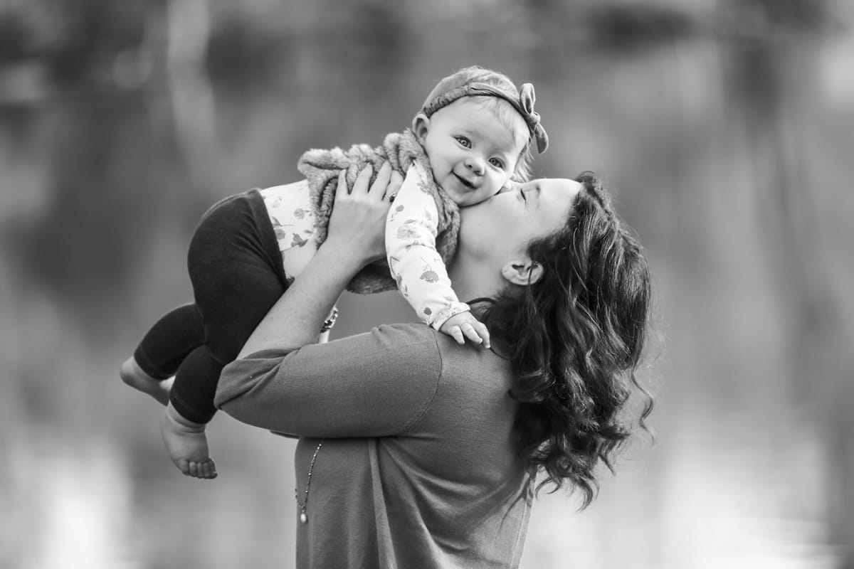 A black and white portrait of a woman lifting a baby in the air and kissing her.