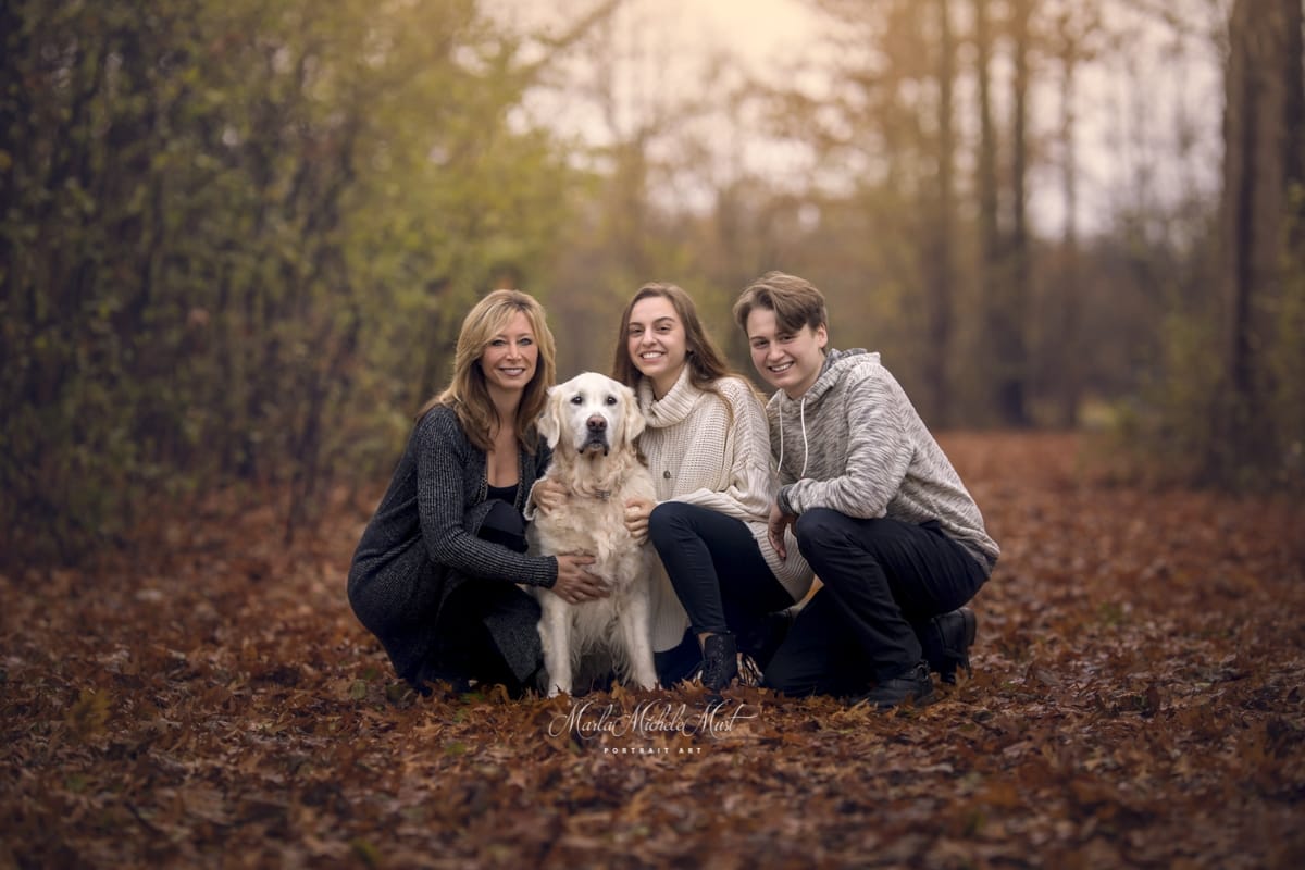 A family of three in fall attire pose for a portrait with their white dog in the forest.
