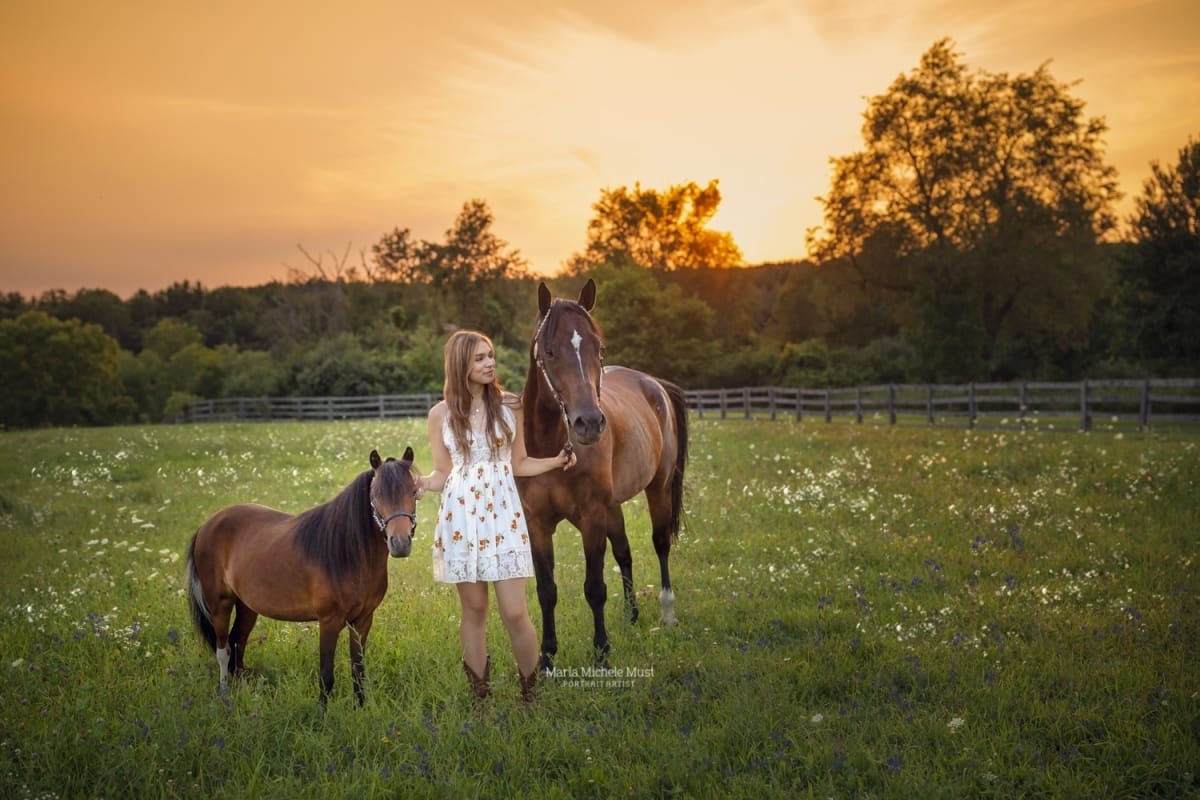 An equine senior photoshoot on a Detroit, Michigan-area farm depicting a girl walking through a field with her two horses at golden hour.