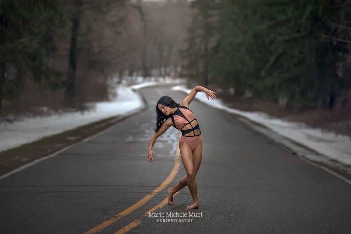 Dancer gracefully embodies a lyrical pose, gracefully extending her arms long and reaching outwards and upwards above her, captured by a talented Detroit photographer on a snowy Michigan road..