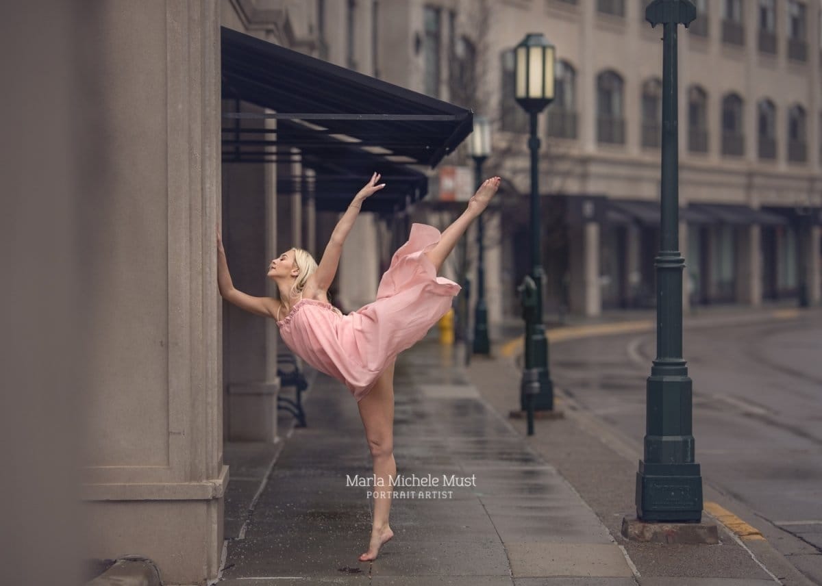 The elegance of a dancer's penché, beautifully graceful in a light pink dress on a rainy city street, photographed during a dance photoshoot by a Detroit photographer.