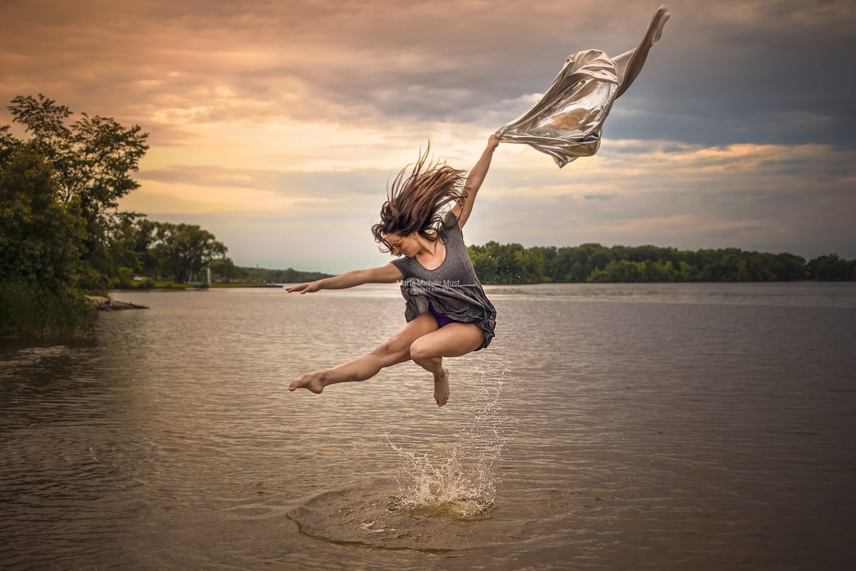 A Detroit dancer captures the essence of grace in a stunning leap above water while holding a silver, flowing cloth during a dance photoshoot.