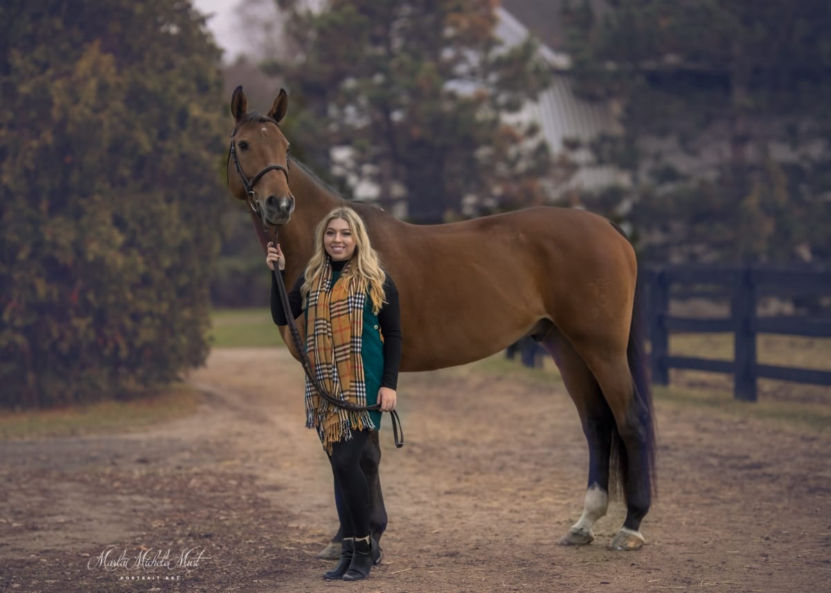 A memorable autumn photograph by a Detroit equine photographer, illustrating the heartwarming bond between a horse and its owner standing proudly next to each other as she hold the reigns and smiles.