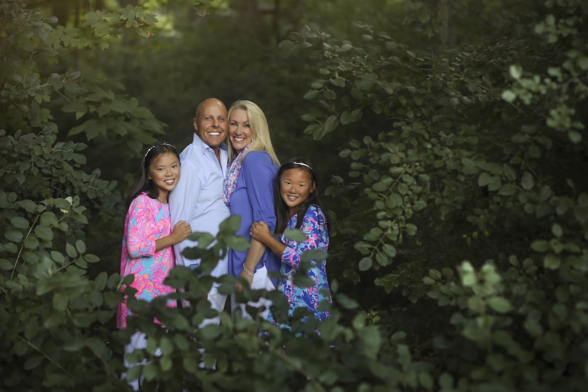 A portrait of a family of four smiling and standing among trees and bushes.