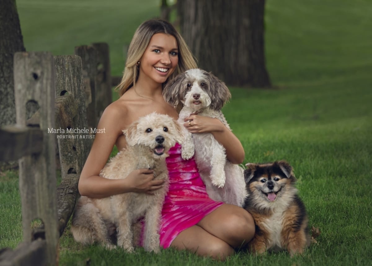 A high school senior's portrait exudes charm and poise as she poses with three dogs in a grassy yard, taken by a skilled Detroit photographer during a senior photoshoot.