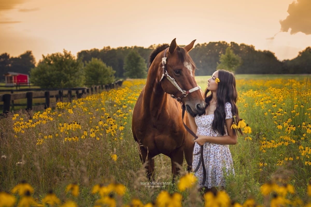 In this high school senior portrait, a girll reaches up to pet her horse as they stand in a field of flowers near a Michigan forest.
