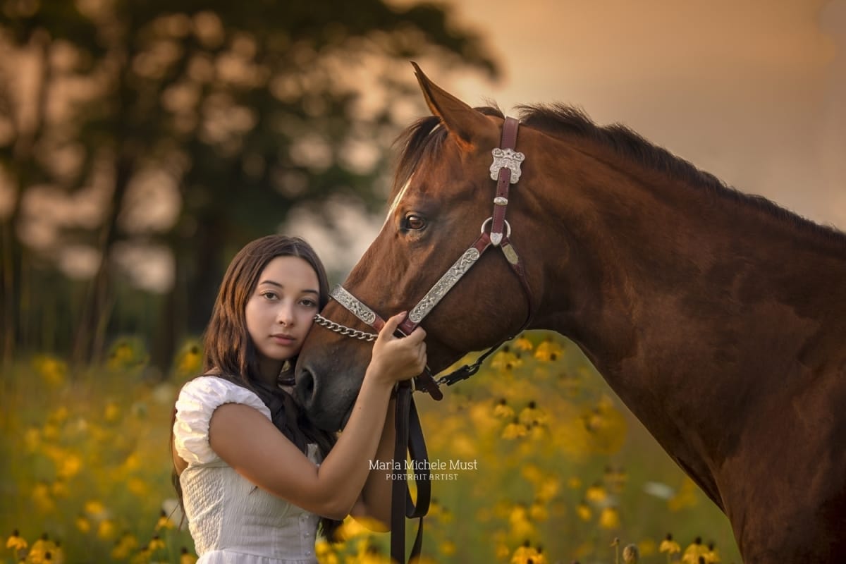 Horse owner portrait for a Detroit equine photographer: Embracing the horse's nose with care in a field of yellow wildflowers, a tree and golden sky behind them.