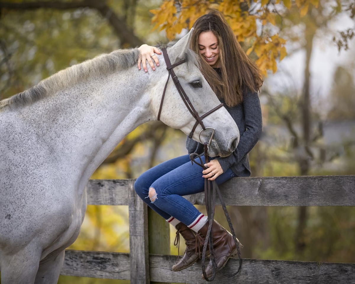 Detroit equine photographer's portrait: Horse owner gently grasping the horse's nose while sitting on a white fence against a background of golden-leaved trees.