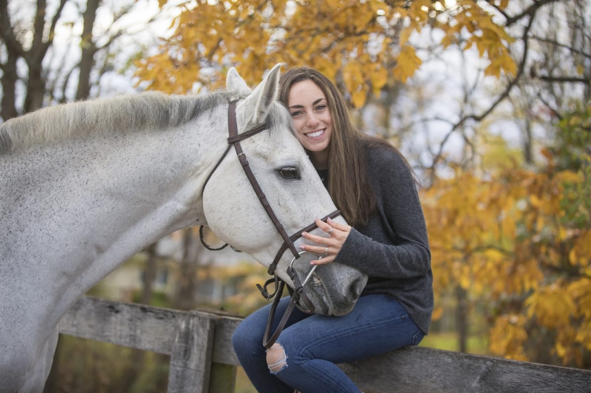 Detroit equine photographer's portrait: Horse owner gently grasping the horse's nose while sitting on a white fence against a background of golden-leaved trees.