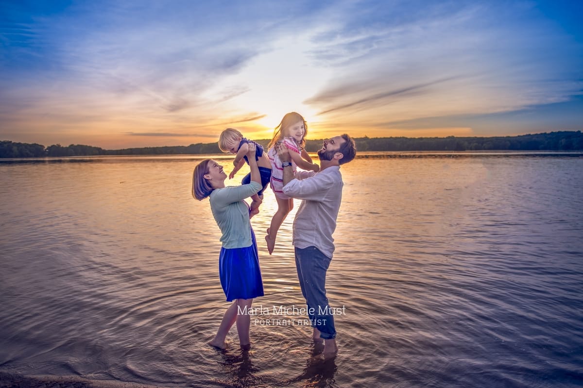 Local Detroit photographer captures a family portrait of a couple holding up each of their children in the air while standing on the beach at sunset.