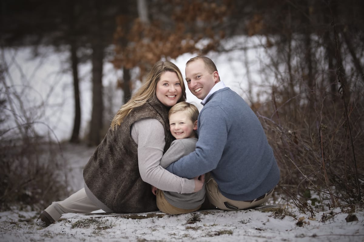 A husband and wife sit by a snowy riverbank with their daughter as they look over their shoulder for a family photo.