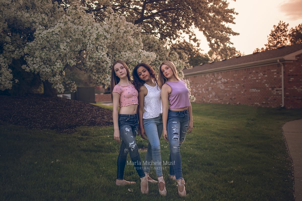 Three dancers in casual clothes pose shoulder to shoulder while extending their legs to showcase matching pointe shoes during a Detroit dance portrait photoshoot.