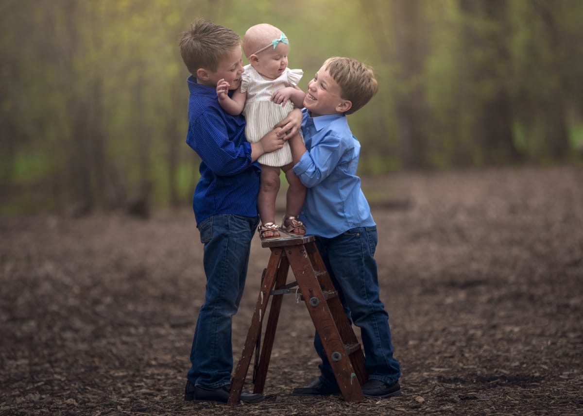 Two young boys embrace their infant sibling that is standing on a small ladder.