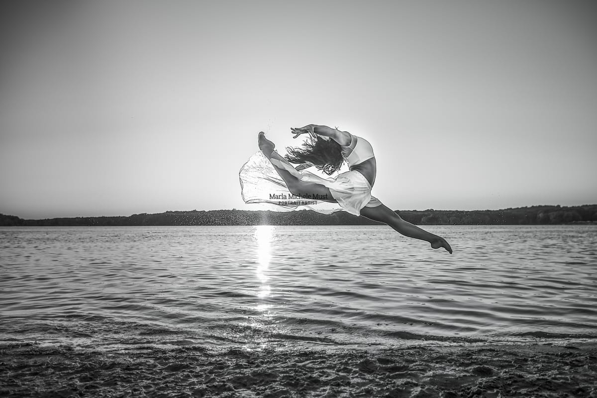 Black and white photo of a dancer on a Michigan beach gracefully ascending into a firebird leap, skillfully captured by a Detroit-based photographer.
