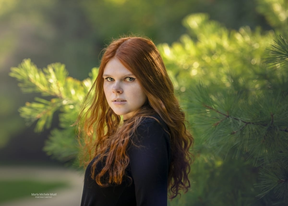 A high school senior portrait photoshoot captures the moment when a girl looks into the distance against a vibrant background of pine branches
