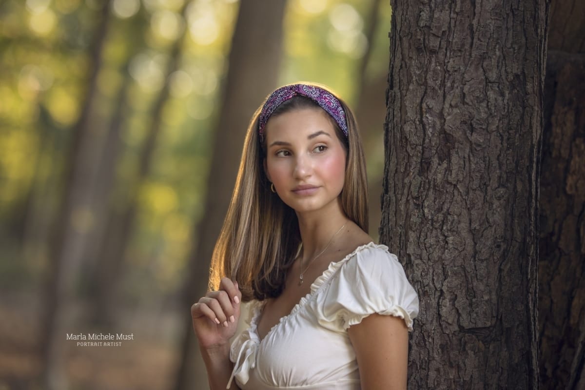 A high school senior portrait photoshoot captures the moment when a girl looks into the distance while leaning against a tree in a fantasy inspired outfit