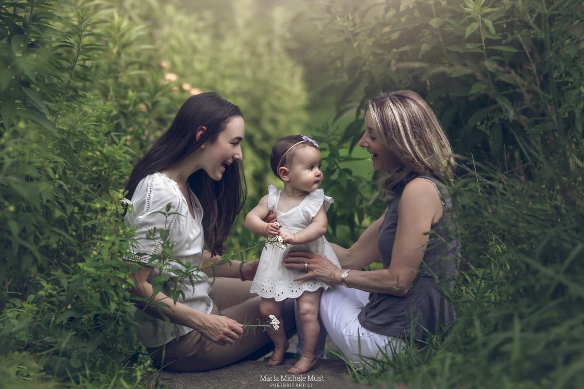 Two women sit with a young toddler aged girl on a bed of grass on a wooded trail somewhere near Detroit.