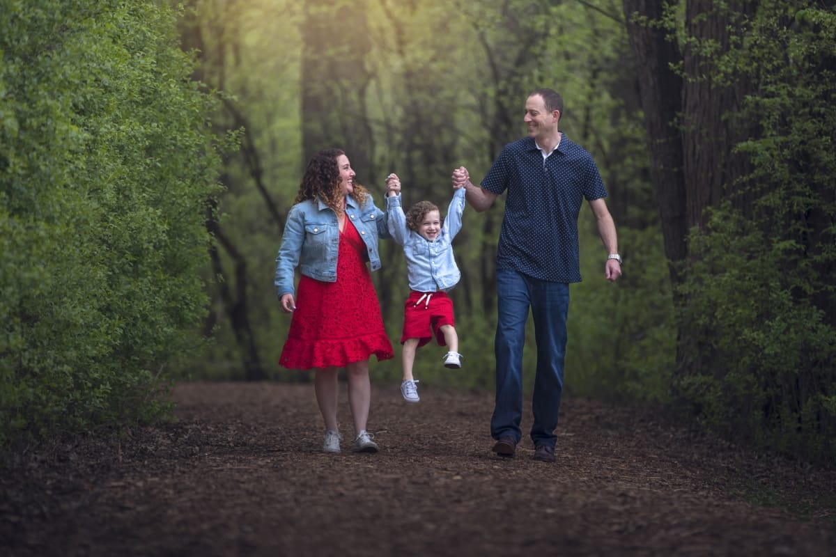 A family photoshoot where a mother and daughter wear red dresses and jean jackets while walking with their father wearing black clothing. They hold her hands and swing her in the air.