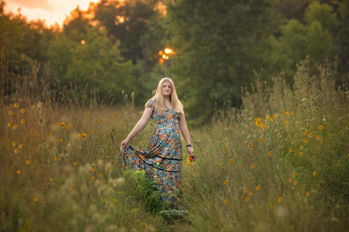 A high school senior portrait photoshoot captures the moment when a girl holds the edge of her long floral dress while walking through a field of wildflowers towards the camera