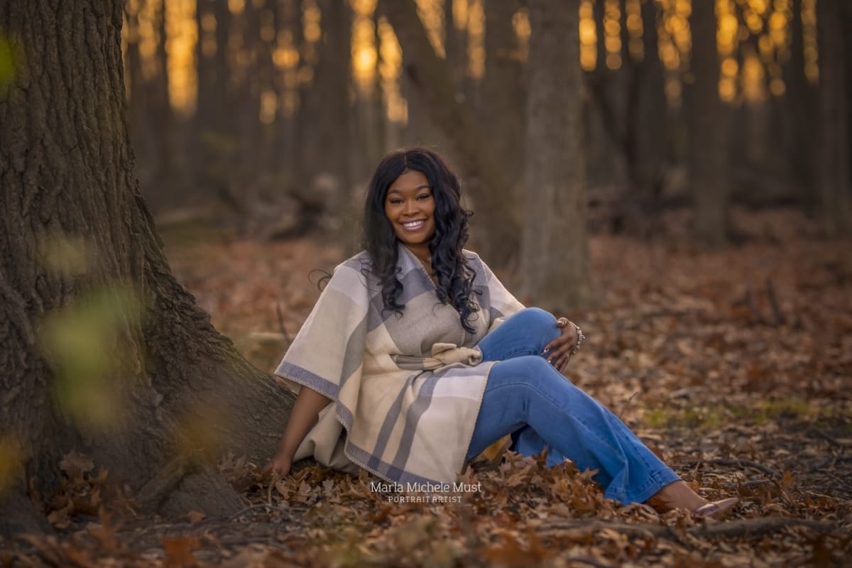 A high school senior portrait photoshoot captures the moment when a girl in cozy fall apparel sits on a bed of autumn leaves in a Detroit forest.