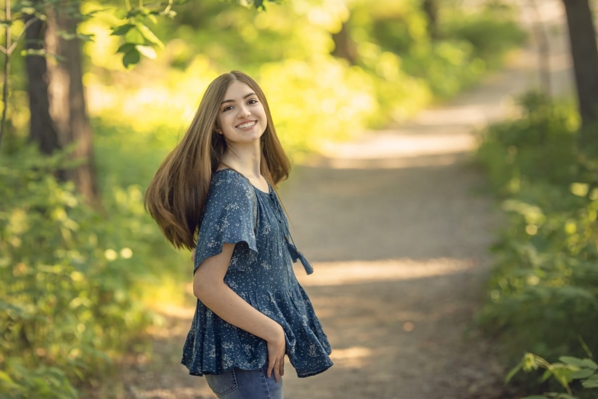 A high school senior portrait photoshoot captures the moment when a girl smiles over her shoulder while twirling on a Detroit forest trail