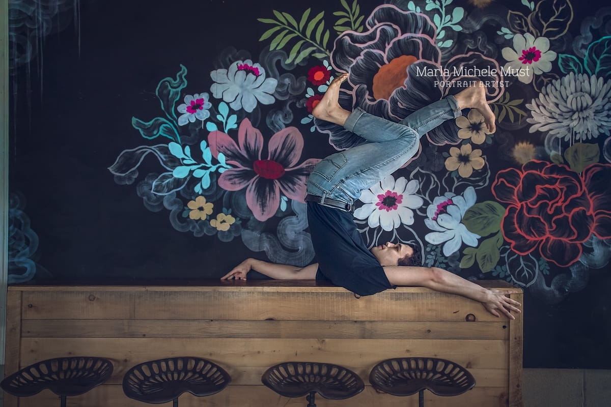 Dancer poses on his back in profile, arms contemporarily posed near his head, legs in a bicycle position above him against a mural depicting a bright floral arrangement during a dancer photoshoot.