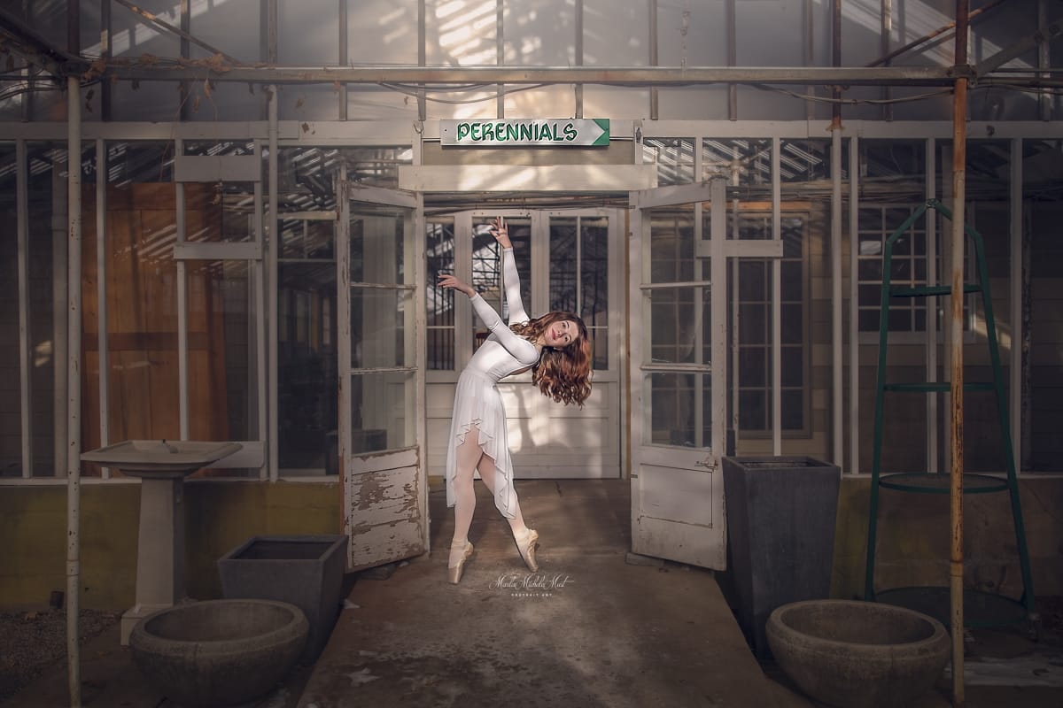 Dancer gracefully poses near a doorway while wearing an ethereal white dress in this captivating image from a dancer photoshoot with a talented Detroit photographer in a Michigan flower nursery.