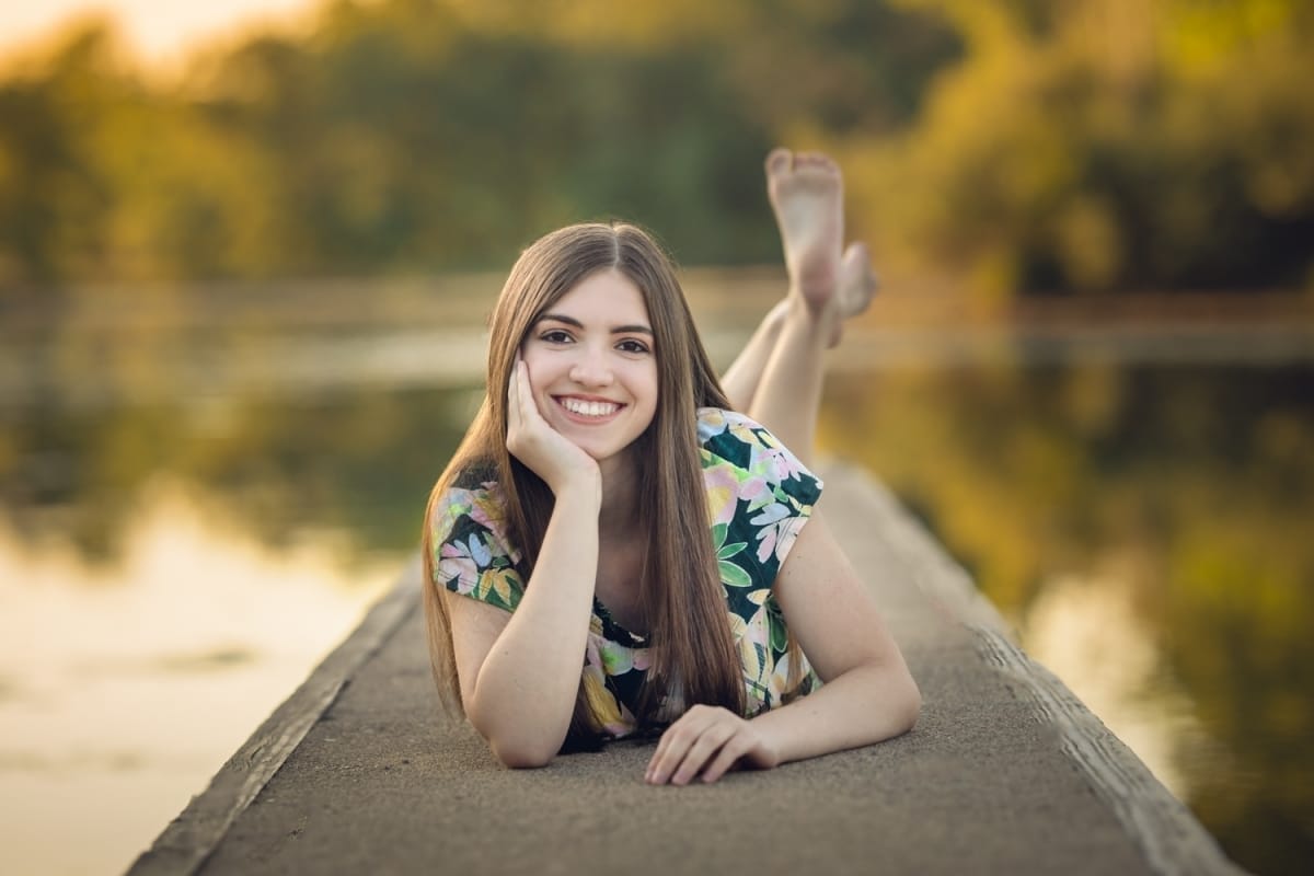 A high school senior portrait photoshoot captures the moment when a girl smiles at the camera, hand resting on her chin, as she lays on the dock of a Detroit park pond.
