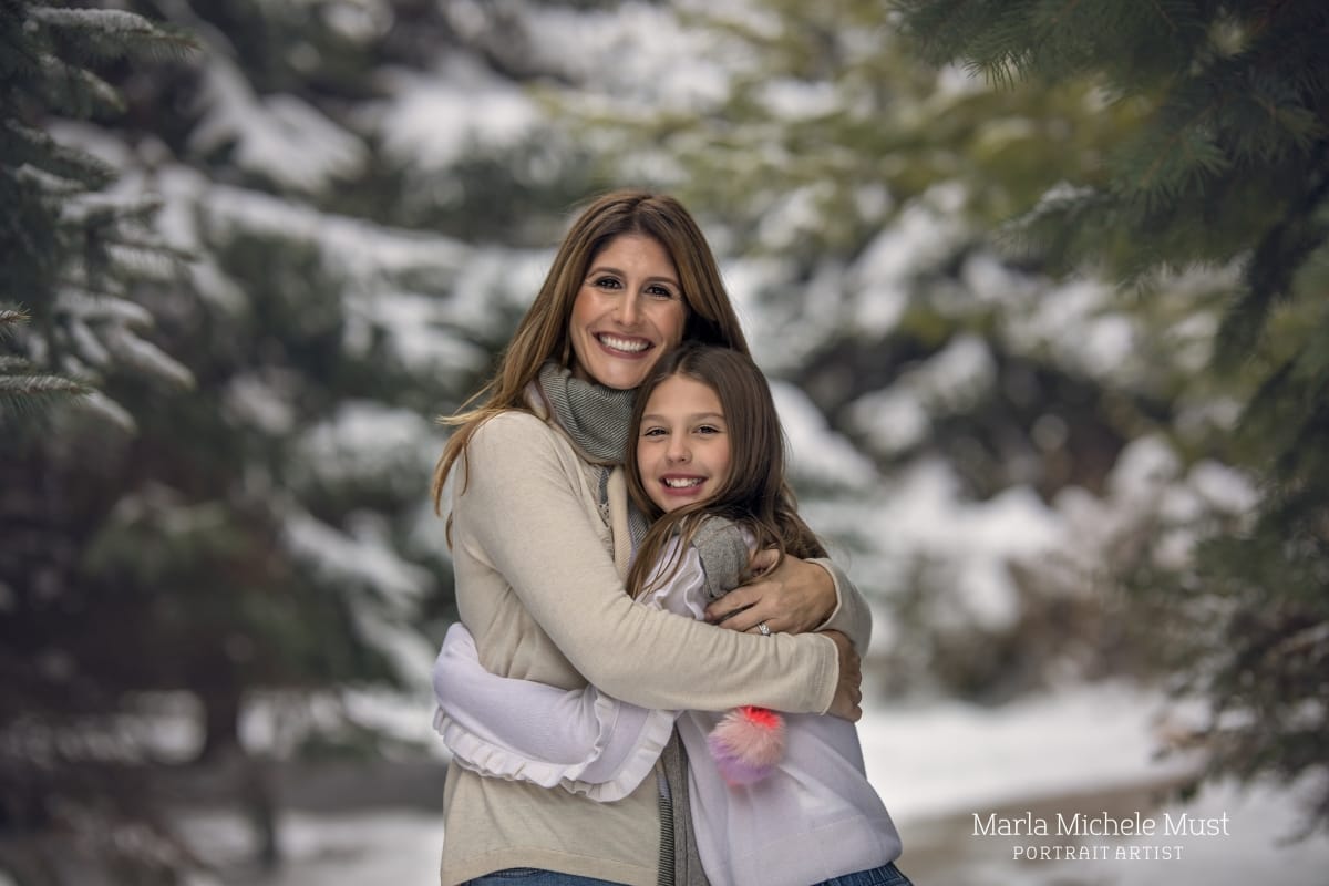 A mother holds her daughter close before a snowy mountainside for a mother-daughter photoshoot