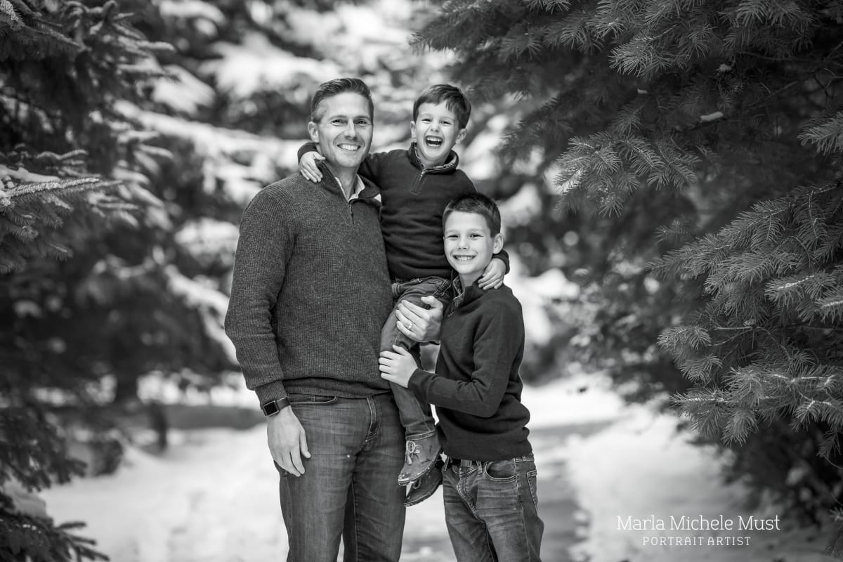black and white family photoshoot A father and his two young sons stand towards the camera before a snowy mountainside.