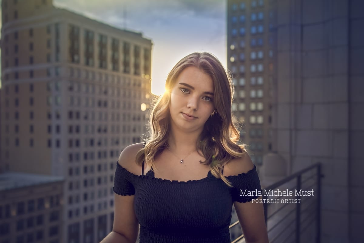 A classic senior portrait of a girl looking into the camera with a city at sunset behind her